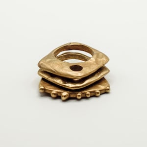 Puede incluir: Un conjunto de tres anillos apilables de color dorado con un acabado texturizado martillado. Los anillos están apilados uno encima del otro, con el anillo inferior que tiene una fila de pequeñas cuentas alrededor del borde.