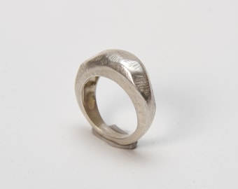 May include: A silver ring with a textured, geometric design. The ring is made of a single piece of metal and has a unique, modern look.