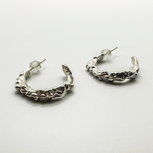May include: A pair of silver-colored hoop earrings. The earrings have a textured, organic design with irregular bumps and a post closure. The earrings are displayed on a white background.