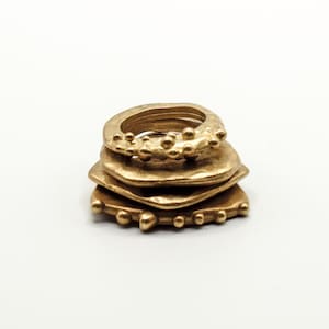 Può includere: Un set di quattro anelli dorati impilati uno sopra l'altro. Gli anelli sono di forme e dimensioni diverse, e l'anello superiore ha un design testurizzato.