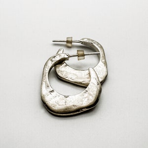 May include: A pair of silver-toned, hammered-texture hoop earrings. The earrings have a square shape with rounded corners and post-style closures. The earrings are displayed on a white background.