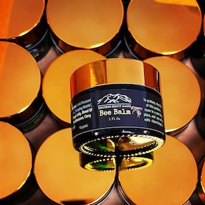 May include: A gold jar of Bee Balm with a black label. The label says "Northern Beauty Alaska" and "Bee Balm" with a bee and mountain graphic. The jar is surrounded by other gold jars.