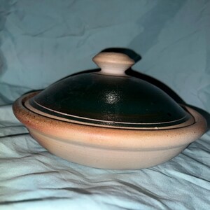 May include: A round, shallow ceramic lidded dish featuring a natural, unglazed terracotta-colored base and a contrasting dark, glossy forest green glazed lid with a central knob handle, showcasing a smooth, rounded silhouette and earthy tones.