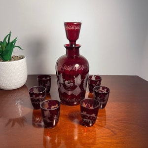 May include: A set of seven ruby red glass decanter and shot glasses. The decanter has a stopper and is etched with a white design of a deer and trees. The shot glasses are also etched with a white design.