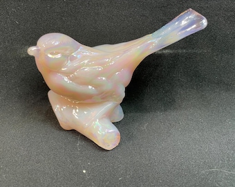 Puede incluir: Una figurita de pájaro de vidrio iridiscente rosa posada en una rama. El pájaro tiene una cola larga y está detallada con plumas intrincadas.