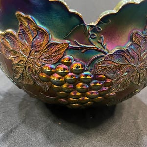Puede incluir: Cuenco de vidrio iridiscente con dise&ntilde;o de uvas y hojas. El cuenco tiene un borde festoneado y una rica paleta de colores p&uacute;rpura, dorado y verde. Las uvas y las hojas est&aacute;n en relieve, a&ntilde;adiendo textura.
