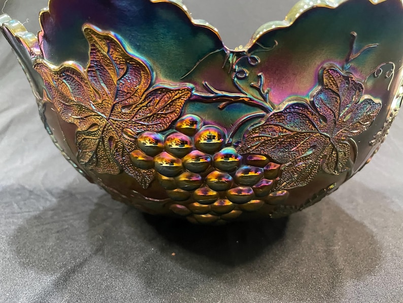 Puede incluir: Cuenco de vidrio iridiscente con dise&ntilde;o de uvas y hojas. El cuenco tiene un borde festoneado y una rica paleta de colores p&uacute;rpura, dorado y verde. Las uvas y las hojas est&aacute;n en relieve, a&ntilde;adiendo textura.