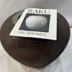 May include: A dark brown Raku pottery vessel with a rounded shape. A white label with the word "RAKU" and "LES MITCHELL" is attached. The label features a black and white image of a cracked pottery design.