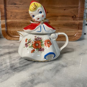 May include: A vintage ceramic teapot with a red and white floral design. The lid features a girl with blonde hair wearing a red hooded cloak.
