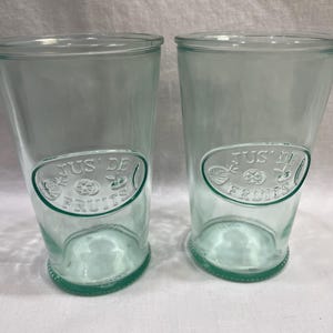 May include: Two clear, light green glass tumblers. Each tumbler has a rounded base and a slightly flared top. An oval emblem with the words "JUS DE FRUITS" is embossed on the front of each glass. The glasses are ideal for serving juice or other beverages.