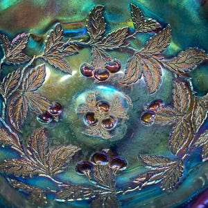 Puede incluir: Primer plano de un cuenco de vidrio iridiscente con un dise&ntilde;o en relieve detallado. El cuenco presenta un patr&oacute;n de hojas y cerezas en tonos dorados, verdes y morados. La superficie del cuenco refleja la luz, creando un efecto brillante.