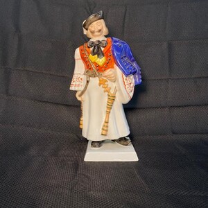 May include: A porcelain figurine of a man wearing a white and red patterned shirt, a blue jacket, and a black hat. He is holding a staff in his right hand and has a white beard and mustache.