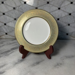 May include: A green and gold decorative plate with a white center. The plate is on a wooden stand.