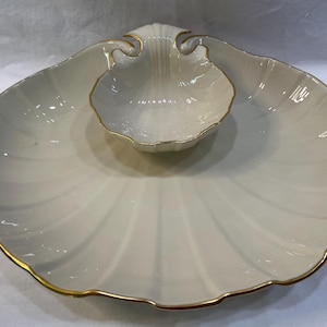 May include: A two-piece serving dish set in a seashell design. The set includes a large, scalloped platter and a smaller, shell-shaped bowl, both with a creamy white finish and gold trim. Ideal for serving appetizers or snacks.