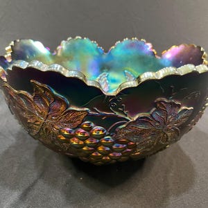 Puede incluir: Cuenco de vidrio iridiscente con dise&ntilde;o de uvas y hojas. El cuenco tiene un borde festoneado y un acabado oscuro multicolor con tonos de morado, verde y dorado. Las uvas y las hojas est&aacute;n en relieve en el exterior.