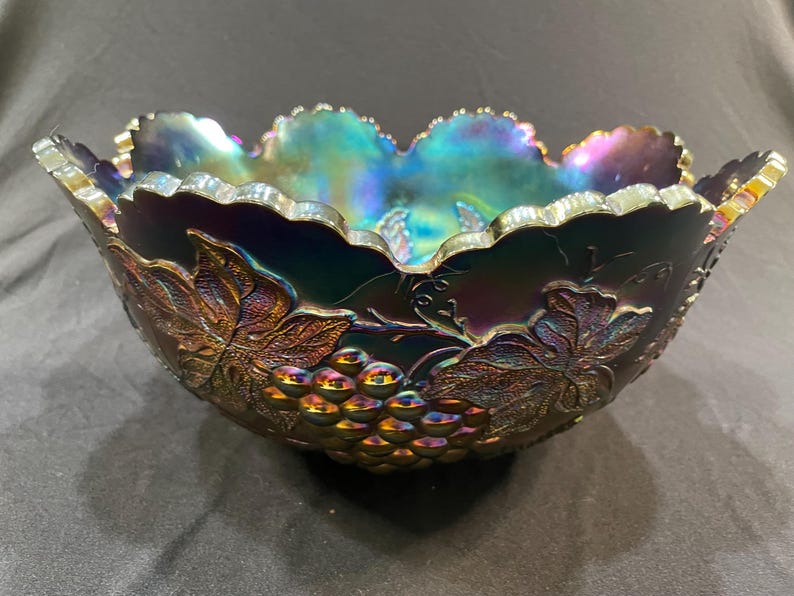 Puede incluir: Cuenco de vidrio iridiscente con dise&ntilde;o de uvas y hojas. El cuenco tiene un borde festoneado y un acabado oscuro multicolor con tonos de morado, verde y dorado. Las uvas y las hojas est&aacute;n en relieve en el exterior.