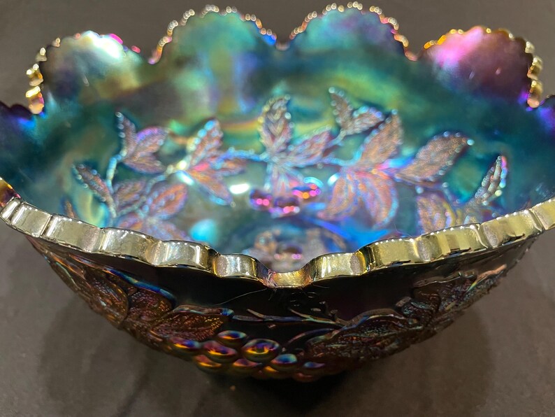 Puede incluir: Un cuenco de cristal de carnaval iridiscente con borde festoneado. El cuenco presenta un dise&ntilde;o de uvas y hojas en tonos morados, azules y dorados. El borde est&aacute; adornado con una banda dorada, a&ntilde;adiendo un toque de elegancia a la pieza vintage.