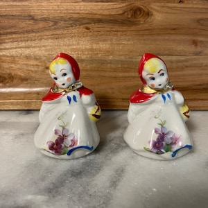 May include: A pair of ceramic salt and pepper shakers shaped like little girls wearing red hooded cloaks. The girls have blonde hair, blue eyes, and are holding baskets. The shakers are decorated with floral designs in shades of purple, pink, and green.