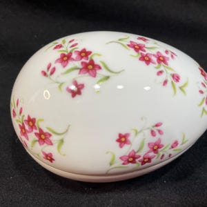 May include: White porcelain egg-shaped trinket box with a floral design featuring pink flowers and green leaves.