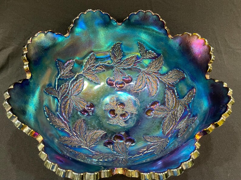 Puede incluir: Cuenco de vidrio azul iridiscente con borde ondulado y dise&ntilde;o de cerezas y hojas. El cuenco tiene un borde festoneado con ribete dorado. La superficie del cuenco refleja una gama de colores, incluyendo azul, verde y morado.