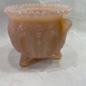 May include: A peach-colored, footed glass bowl with a beaded rim and ornate embossed detailing. The bowl has three small feet and a rounded body. The glass has a slight iridescence, giving it a soft, glowing appearance. A decorative piece.