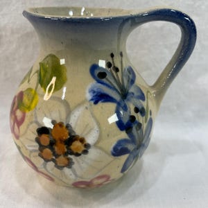 May include: A small, hand-painted ceramic pitcher with a rounded body and a curved handle. The pitcher is beige with blue trim and is decorated with colorful floral designs in blue, white, pink, yellow, and brown.