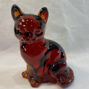 May include: A glossy ceramic cat figurine in shades of red, black, and orange. The cat is in a seated position with its ears perked up. The figurine has a smooth, reflective surface.