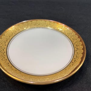 May include: A small, white saucer with a gold rim and intricate floral pattern. The saucer is made of porcelain and has a glossy finish.