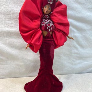 May include: A fashion doll in a long, dark red velvet gown with a matching headpiece and bodice, adorned with sparkling red gems. The doll has brown hair and is wearing earrings. Large, red fabric wings extend from the doll's shoulders.