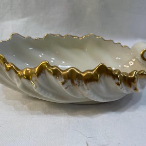 May include: A white, shell-shaped dish with a gold-painted rim. The dish has a wavy, textured design and a curled accent on one end. The gold trim adds a touch of elegance to this decorative piece.