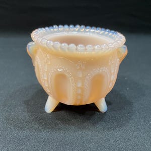May include: A small, pink, glass cauldron with a beaded rim and three legs. The cauldron has a decorative design of arches and dots.