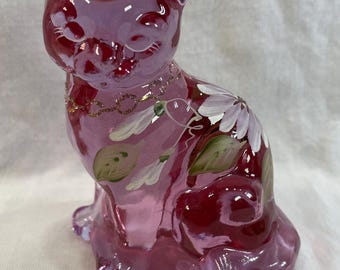 May include: A pink glass cat figurine with painted white flowers and green leaves. The cat has a gold-colored collar and is sitting upright. The figurine is a decorative item.