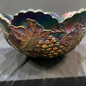 Puede incluir: Un cuenco de vidrio iridiscente con un dise&ntilde;o de uvas y hojas. El cuenco tiene un borde festoneado y un acabado oscuro multicolor con tonos de morado, azul y dorado. El cuenco probablemente se utiliza para servir.