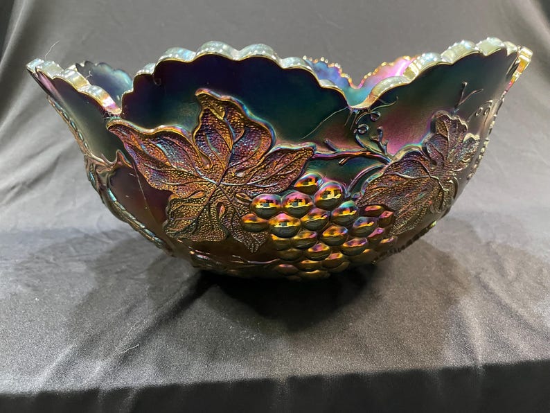 Puede incluir: Un cuenco de vidrio iridiscente con un dise&ntilde;o de uvas y hojas. El cuenco tiene un borde festoneado y un acabado oscuro multicolor con tonos de morado, azul y dorado. El cuenco probablemente se utiliza para servir.