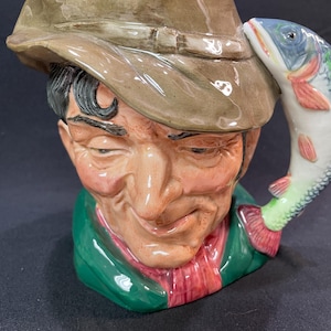 May include: A ceramic pitcher shaped like a man's head wearing a brown hat with a fish handle. The man has a mustache and a sly smile. The fish is painted in shades of blue, green, and red.