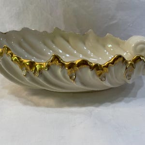 May include: Cream-colored, seashell-shaped decorative bowl with a gold-painted rim. The bowl has a spiral design on one end and a wavy, textured edge. The gold trim adds a touch of elegance to this decorative piece.