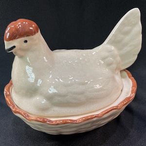 May include: A white ceramic chicken with a brown head and a brown nest. The chicken is sitting in the nest with its tail feathers sticking up.