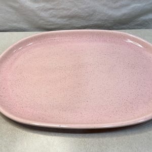 Peut inclure: Un plat ovale en céramique rose clair avec une surface mouchetée. Le plat a un bord légèrement surélevé et une finition lisse et brillante. Ce plat est adapté pour servir des aliments.