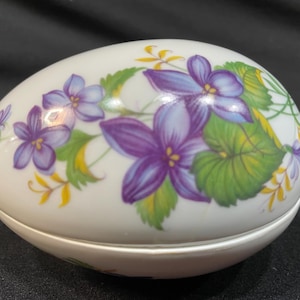 May include: A white ceramic egg-shaped trinket box with a floral design. The box is decorated with purple and blue flowers and green leaves.