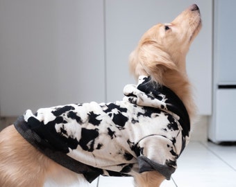 May include: A light brown dog wearing a black and white tie-dye hoodie. The dog is standing on a white tiled floor and looking up.