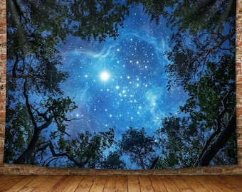 May include: A tapestry depicting a night sky scene. The image features a vibrant blue sky filled with numerous stars and a bright central light source. Dark green tree branches frame the celestial view, creating a natural border. The tapestry hangs against a brick wall.