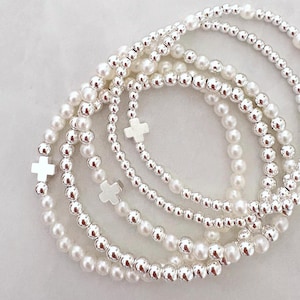 May include: A selection of beaded bracelets. The bracelets combine silver-coloured beads and white pearls. Some bracelets include small, silver cross charms. The bracelets are arranged on a white surface.