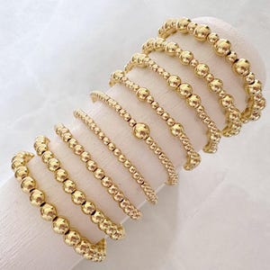 Könnte beinhalten: Eine Sammlung goldener Perlenarmbänder in verschiedenen Größen und Designs. Die Armbänder sind auf einer weißen zylindrischen Form vor einem weichen, weißen Hintergrund ausgestellt. Die Perlen sind rund und gleichmäßig verteilt.