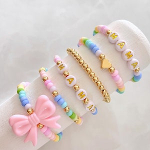 May include: A set of five colorful beaded bracelets with gold accents. The bracelets feature the word "MAMA" spelled out in white beads with gold lettering. One bracelet has a gold heart charm. One bracelet has a pink bow charm.