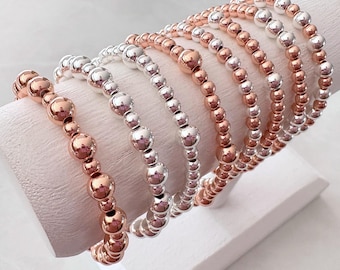 May include: A display of beaded bracelets in rose gold and silver tones. The bracelets are made of small, round beads and are arranged on a white, angled display stand. The bracelets are a mix of solid rose gold and silver, creating a contrast.