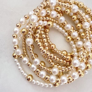以下が含まれることがあります： 金色のビーズブレスレットとパールのブレスレットのコレクション。ブレスレットは、金色のビーズと白いパールの組み合わせです。ブレスレットは重ねられており、豪華でエレガントな外観を作り出しています。