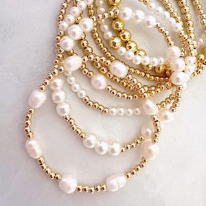 May include: A collection of gold beaded bracelets and pearl bracelets. The bracelets are in various sizes and styles, with a mix of small gold beads and larger pearls. The jewelry is arranged in a layered, stacked fashion.
