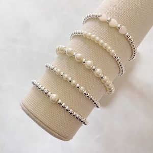 Op de afbeelding: Vijf zilveren kralen armbanden met witte parel accenten. De bovenste armband heeft twee witte hartvormige kralen.