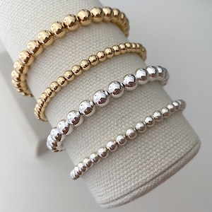 May include: Four beaded bracelets stacked on a fabric display stand. The top two bracelets are gold, and the bottom two are silver.