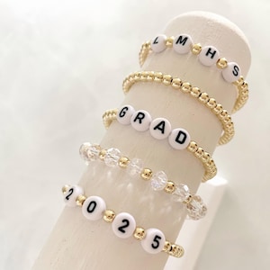 May include: Five gold beaded bracelets with white letter beads spelling out "GRAD", "2025", and "MHS".